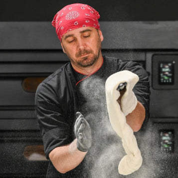Man in a black shirt and red bandana holding a ball of dough in front of a pizza oven.
